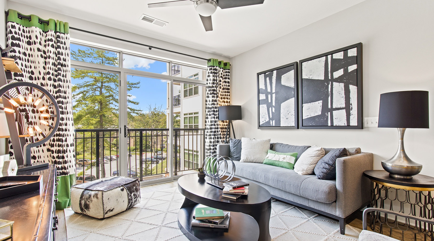 A living room with a large window and balcony at Vista Wilde Lake, featuring a gray couch, modern decor, patterned curtains, a black coffee table, and accent lighting